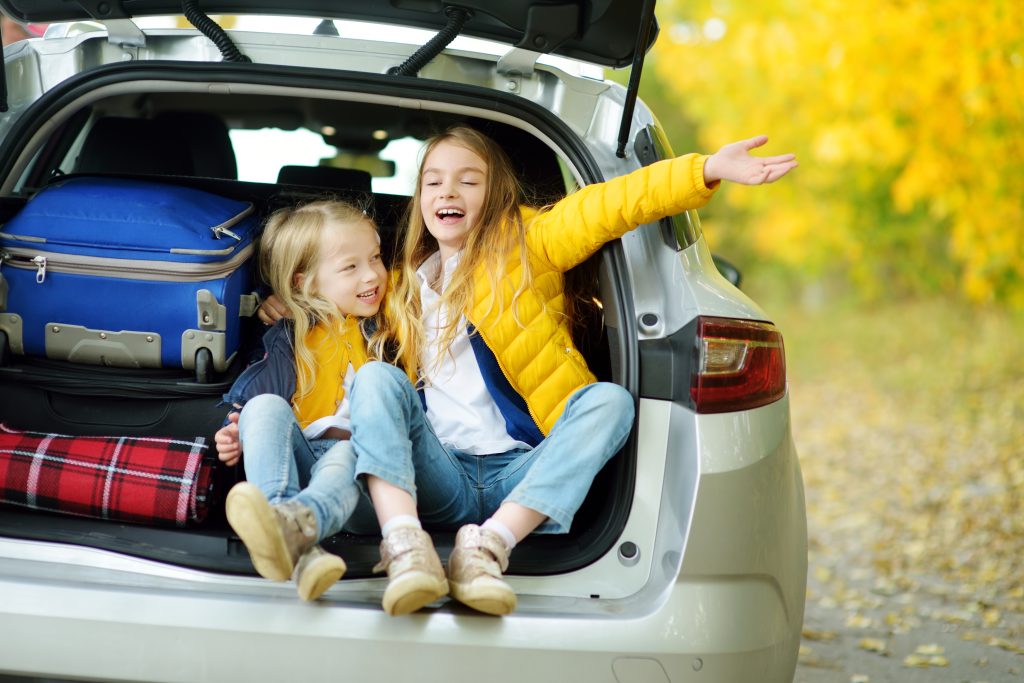 two little girls giggling sitting in the trunk of the car next to suitcases packed